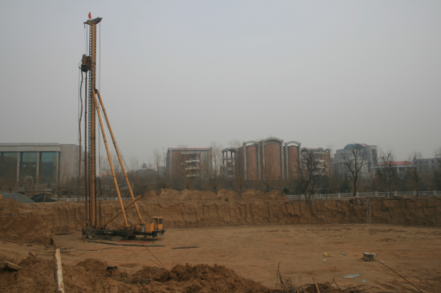 Eine Baustelle mit einem Kran in der Mitte, umgeben von Gebäuden, Bäumen und einem klaren blauen Himmel, mit verstreuten Gegenständen auf dem bodenbedeckten Grund.