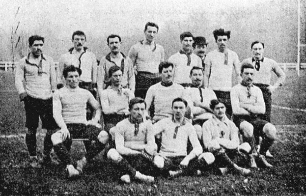 Ein Schwarz-Weiß-Foto einer Fussballmannschaft aus den 1920er Jahren, mit einigen Spielern stehend und anderen auf dem Boden sitzend, vor einer Kulisse aus Pfählen, Bäumen und einem klaren Himmel.