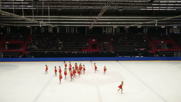 Eine Gruppe von Menschen, die auf einer Eisbahn Schlittschuh laufen, umgeben von einem Publikum, das auf Stühlen sitzt, mit Stangen und Lichtern, die die Bahn von oben beleuchten.