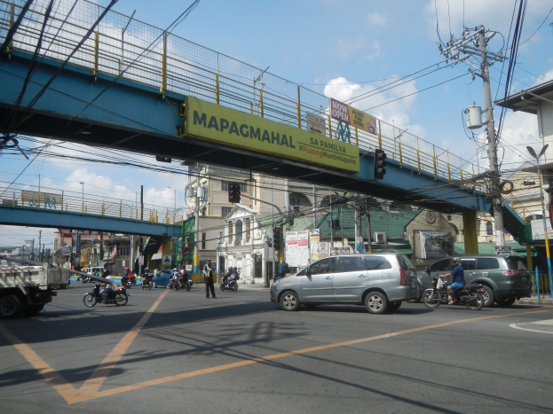 Lebendige Stadtstraße mit Fahrzeugen, Fußgängern, einer Brücke mit Geländern und einem Namensschild, Strommasten mit Drähten, Straßenlaternen, Verkehrsampeln, Gebäuden mit Fenstern und einem bewölkten Himmel.