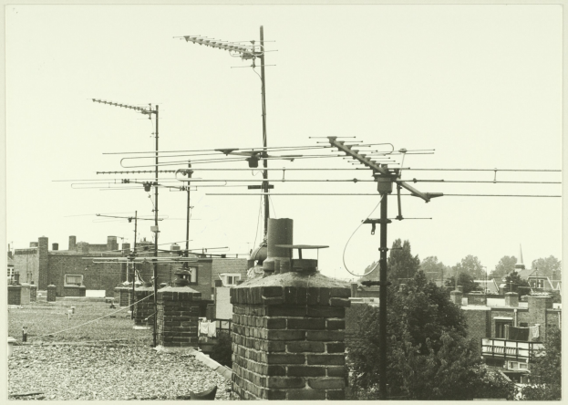 Ein Schwarz-Weiß-Foto einer Stadtlandschaft mit zahlreichen Gebäuden, Bäumen und Fernsehantennen auf einem Dach.