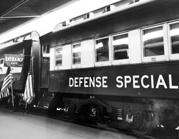 Ein Schwarz-Weiß-Foto eines Zuges mit der Aufschrift 'Defense Special', der auf den Schienen steht, mit zwei Fahnen auf der linken Seite und einer Decke über ihm.