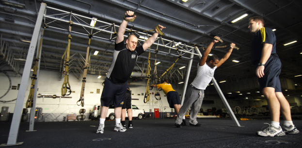 Eine Gruppe von Männern, die Klimmzüge in einem Fitnessstudio mit Metallstangen, Rahmen und Deckenleuchten macht, wobei ein Mann in der Mitte Schuhe trägt.