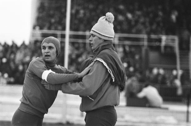 Zwei Männer in Jacken und Mützen kämpfen auf einem Feld mit unscharfen Zuschauern, Pfählen und einem Zaun im Hintergrund, in Schwarz-Weiß.