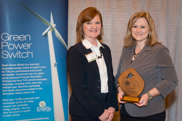 Zwei Frauen stehen in einem Raum, eine hält eine Auszeichnung, mit einem Banner neben ihr und einem Vorhang im Hintergrund.