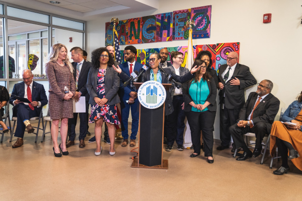 Eine Gruppe von Menschen steht vor einem Podium mit einem Mikrofon und einer Tafel mit dem Text "Sancho City Councilman Joins City Council". Andere sitzen auf Stühlen oder stehen in der Nähe, während auf der Rückwand Flaggen, eine Tafel, ein Feuerlöscher und Deckenleuchten zu sehen sind.