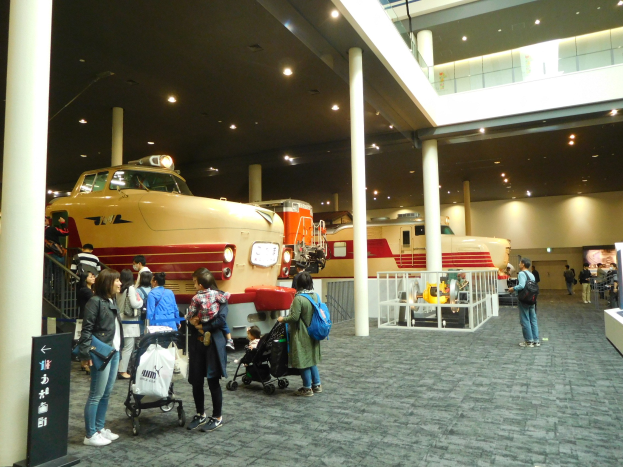 Menschen vor einem Zugmodell in einem Museum mit Informationsschildern, Säulen, Geländern und Deckenleuchten.