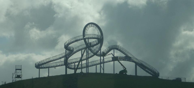 Eine Achterbahn auf dem Höhepunkt eines Hügels mit einem bewölkten Himmel im Hintergrund, umgeben von Stützpfählen und anderen Fahrradelementen.