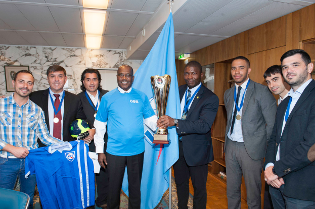 Eine Gruppe von Menschen, die für ein Foto posieren, wobei zwei in der Mitte eine Trophäe halten, einer links ein T-Shirt und ein weiterer links außen einen Ball sowie eine Flagge im Hintergrund.