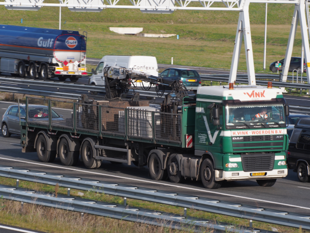 Ein großer Lkw mit einem Anhänger fährt auf einer Straße neben einer Brücke, mit Gras und Geländern auf beiden Seiten und einem klaren blauen Himmel im Hintergrund.