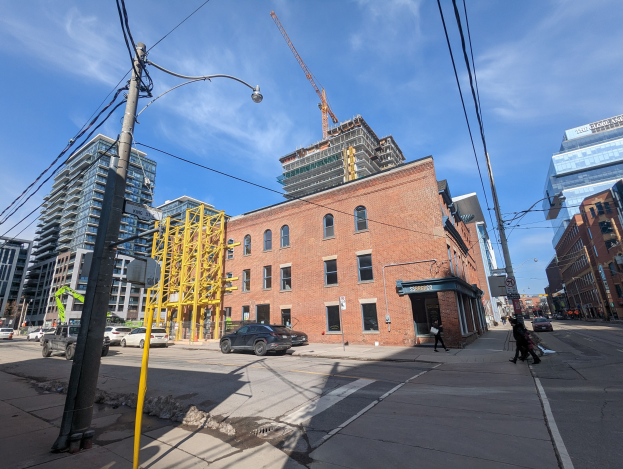 Eine belebte Stadtstraße mit fahrenden Fahrzeugen, Fußgängern auf dem Gehweg, Strommasten mit Kabeln, Straßenlaternen und Gebäuden, einschließlich eines teilweise errichteten Hochhauses mit einem Kran im bewölkten Himmel.