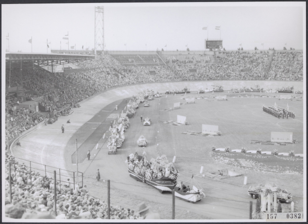 Schwarzes und weißes Foto einer Rennstrecke mit Zuschauern und Fahrzeugen, das Stangen, Flaggen und den Himmel im Hintergrund zeigt.