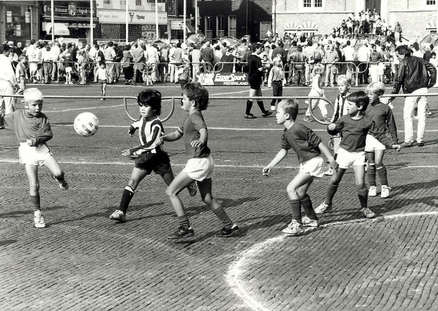 Schwarzes Bild von jungen Jungs, die auf der Straße Fußball spielen, umgeben von einer Zuschauermenge hinter einem Geländer, Gebäude und Pfosten im Hintergrund.