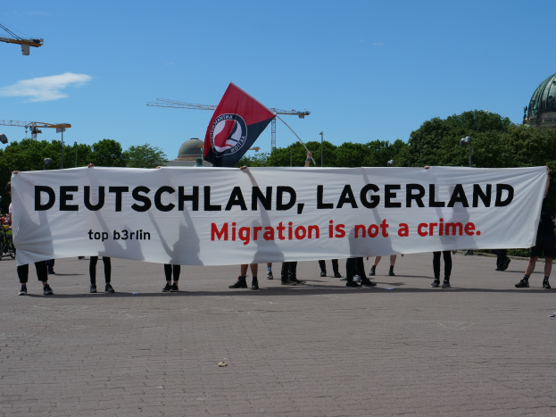 Eine Gruppe von Menschen mit einer Fahne, auf der steht "Deutschland, Lagerland Migration Ist Kein Verbrechen" und einer Flagge, mit Straßenlaternen, Bäumen, Kränen, einem Gebäude und einem bewölkten Himmel im Hintergrund.