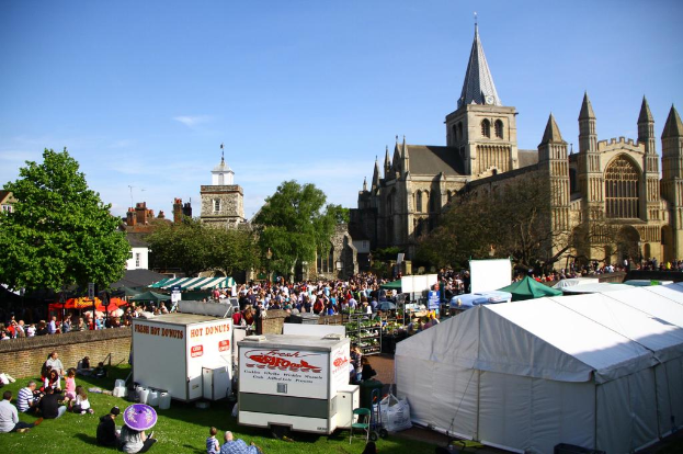Historische Gebäude mit Bäumen davor, ein Turm im Hintergrund, Menschen auf einem Weg mit Geschäften und andere auf der Wiese in der Nähe sitzend.