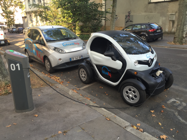 Zwei Elektroautos stehen nebeneinander auf einer Straße, an eine Ladesäule angeschlossen, mit Bäumen und Fenstern im Hintergrund.