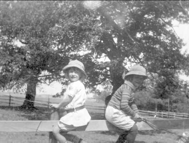 Zwei junge Jungs in Mützen stehen in einem grünen Park und halten Baseballschläger in der Hand, mit Bäumen und einem Zaun im Hintergrund, auf einem Schwarz-Weiß-Bild.