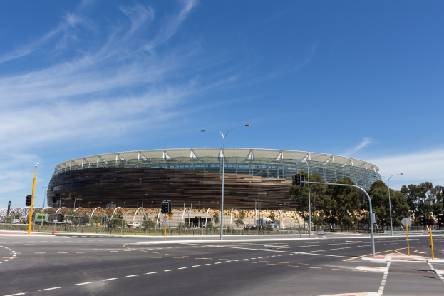 Ein großes Stadion mit starkem Verkehr davor, umgeben von Straßenlaternen, Lichtern, Verkehrsschildern, Geländern, Bäumen und einem bewölkten Himmel.