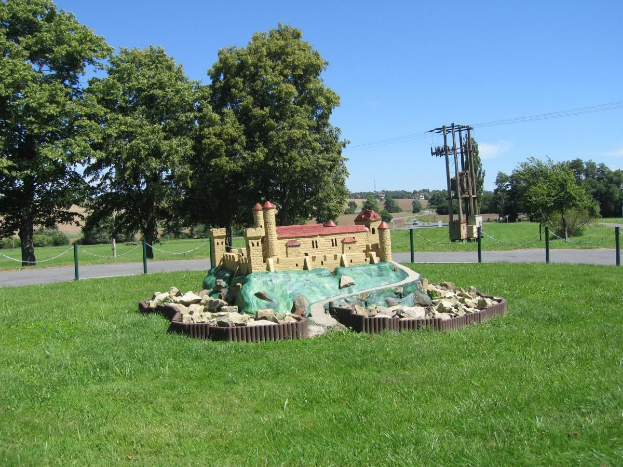 Minigolfplatz mit Rasenfeld, umgeben von einem Zaun mit Pfählen und Ketten, Bäumen, Strommasten mit Drähten und einem bewölkten Himmel im Hintergrund.
