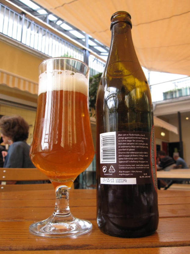 Ein Glas Wein und eine Weinflasche auf einem Tisch mit einer Gruppe von Menschen, einem Zelt, einem Gebäude und Lichtern im Hintergrund.