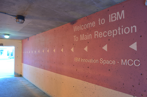 Eine Wand mit "Willkommen bei IBM" beleuchtet von Deckenlampen, mit einer Tür zum IBM Innovation Space MCC im Hintergrund.