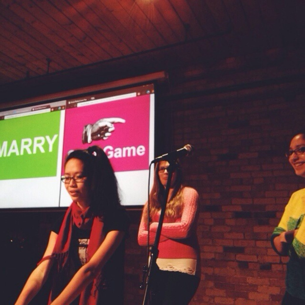 Drei Frauen, die nach links schauen, mit einem Mikrofonständer in der Mitte, einer Wand im Hintergrund und einem Bildschirm mit Text auf der linken Seite.