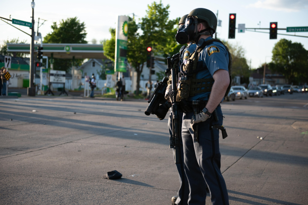 Polizeibeamter in voller Einsatzbekleidung mit einer Waffe an der Straße stehend mit Fahrzeugen, Verkehrszeichen, Schildern, Bäumen, Gebäuden und Himmel im Hintergrund.