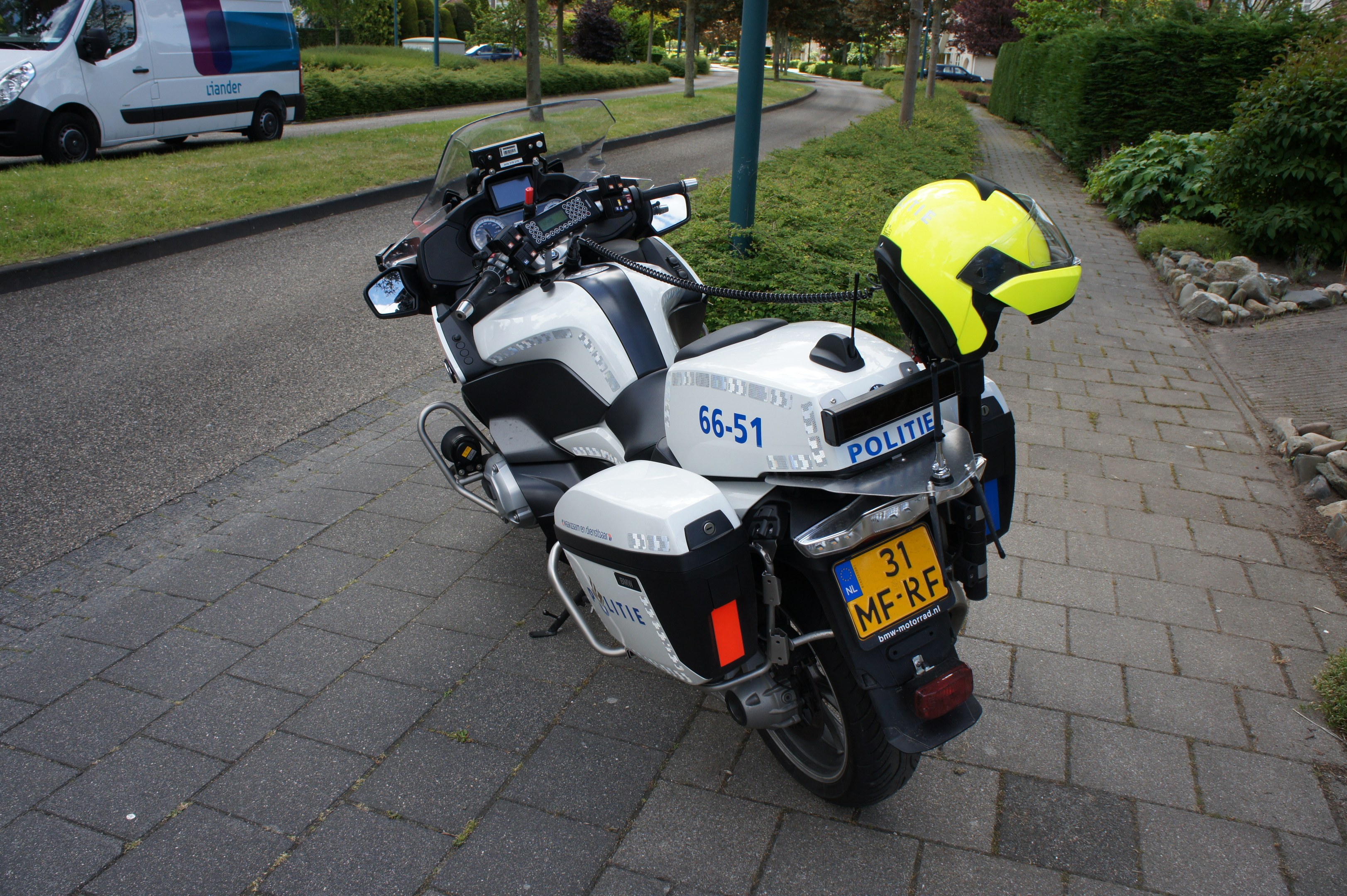 Ein polizeiliches Motorrad am Straßenrand mit einem Helm darauf, umgeben von Bäumen, Pfählen, Gebäuden, Pflanzen, Gras, Steinen und anderen Fahrzeugen.