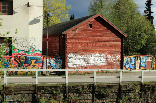 Ein rotes Scheunengebäude mit Graffiti an der Seite, umgeben von einem Zaun, Pflanzen, Gras, einer Straße, einem Lichtmast, einem Gebäude mit Fenstern, Bäumen und einem bewölkten Himmel im Hintergrund.