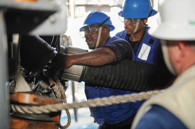 Drei Männer in harten Helmen und Schutzwesten an einer Maschine arbeiten, mit einem Seil im Vordergrund und einem unscharfen Hintergrund.