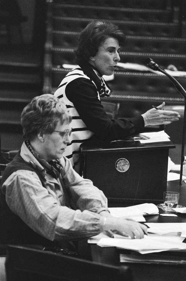 Schwarz-weißes Foto einer Frau, die an einem Tisch im Europäischen Parlament in Strasbourg im Mai 1981 sitzt und einen Stift hält, mit Papieren, einem Glas Wasser und anderen Gegenständen auf dem Tisch, mit Treppen im Hintergrund.