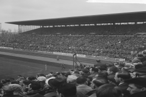 Schwarzes und weißes Foto eines vollen Stadions mit Zuschauern, die ein Fußballspiel verfolgen, mit Bannern, Pfählen, einer Hütte, Bäumen, einem Turm und einem bewölkten Himmel.