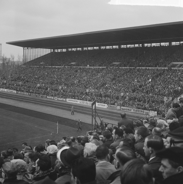 Schwarzes und weißes Foto eines vollen Stadions mit Zuschauern, die ein Fußballspiel verfolgen, mit Bannern, Pfählen, einer Hütte, Bäumen, einem Turm und einem bewölkten Himmel.