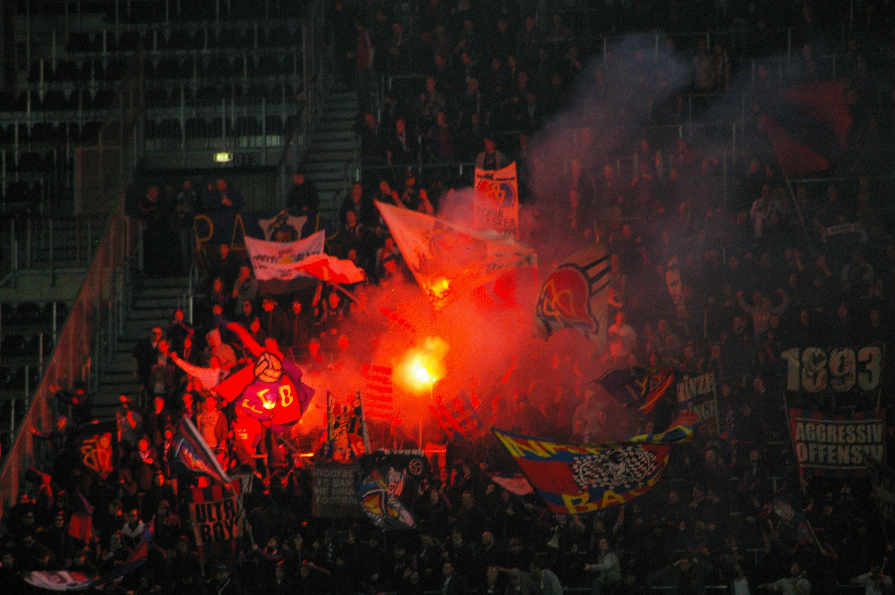 Eine große Menschenmenge in einem Stadion mit Fahnen, Bannern und einem zentralen Feuer, beleuchtet von Scheinwerfern und umgeben von Stufen und Geländern.