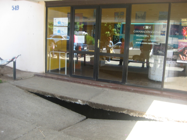 Eine gebrochene Oberfl√§che im Vordergrund mit einer Glast√ľr, Wand und Stange im Hintergrund; die Spiegelung in der Glast√ľr zeigt Fahrzeuge auf der Stra√üe und einige Personen, die stehen.