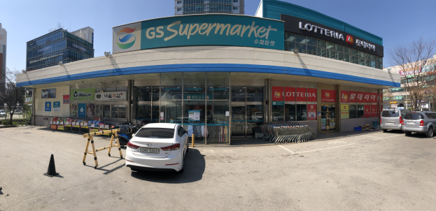 Ein Auto vor einem Laden mit einer "GS Supermarkt"-Aufschrift geparkt, umgeben von anderen Fahrzeugen, Verkehrskegeln und verstreuten Gegenständen, mit Gebäuden, Bäumen und einem klaren blauen Himmel im Hintergrund.