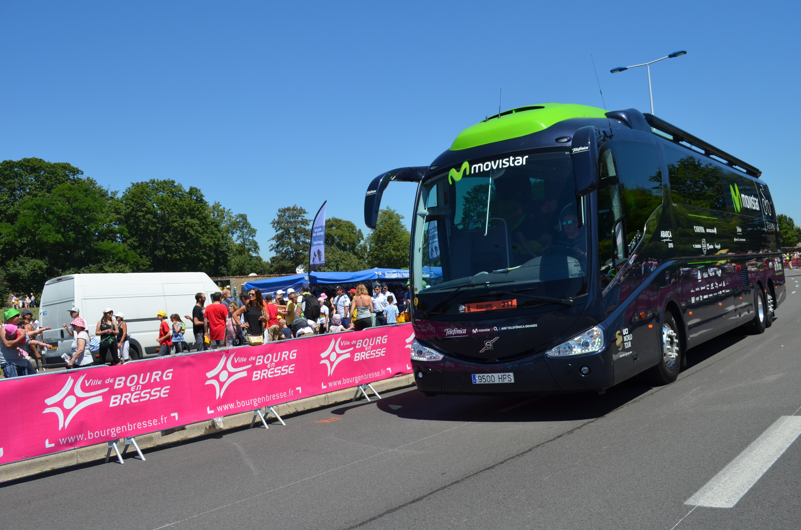 Ein schwarzer und grüner Bus fährt auf einer Straße neben einer Menschenmenge, einige tragen Mützen, mit einem Banner auf der linken Seite, Bäumen und einem klaren blauen Himmel im Hintergrund.