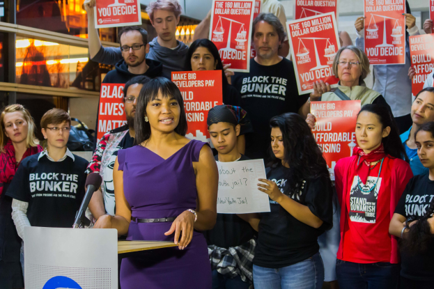 Eine Black-Lives-Matter-Aktivistin spricht an einem Rednerpult mit Mikrofon vor einer Menge mit Schildern, mit Lichtern und Gegenständen im Hintergrund.