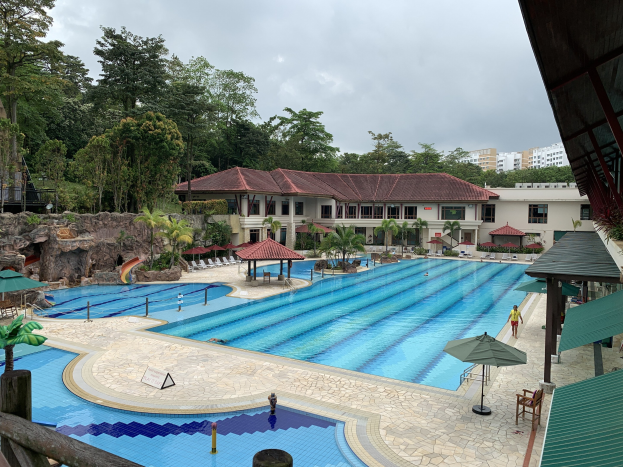 Großes Schwimmbecken im Ritz-Carlton, Kota Kinabalu, mit Menschen, Sonnenschirmen, Liegestühlen, Tischen, Grünpflanzen, Felsen und Gebäuden, einschließlich eines Schuppens auf der rechten Seite und einem sichtbaren Himmel im Hintergrund.