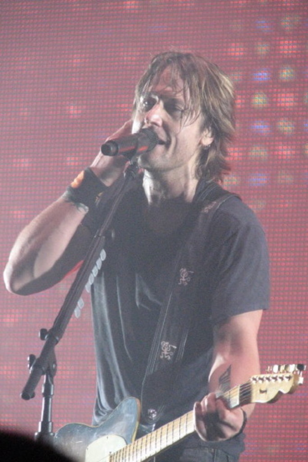 Ein Mann in einem T-Shirt hält eine Gitarre und singt in ein Mikrofon, mit einem Bildschirm oder einer Tafel im Hintergrund.