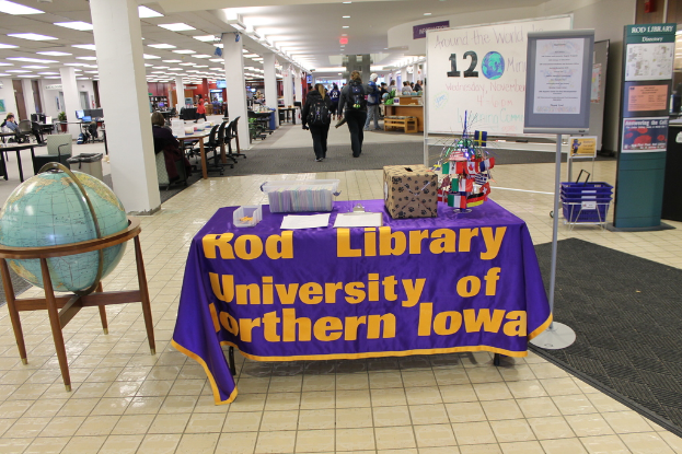 Eine Ausstellung mit einem Tisch, auf dem verschiedene Landesflaggen, eine Kiste, Papiere und eine Fahne liegen, eine Kugel links daneben, ein Anschlagbrett rechts daneben mit einigen Menschen in der Nähe und mehreren Tischen und Stühlen links.