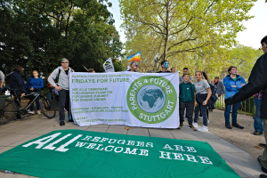 Eine Gruppe von Menschen steht auf dem Boden, einige halten ein Banner mit der Aufschrift "All Refugees Are Welcome Here" und eine Flagge, mit Fahrrädern, einem Zaun, einem Straßenschild, einer Schautafel, Bäumen und einem bewölkten Himmel im Hintergrund.