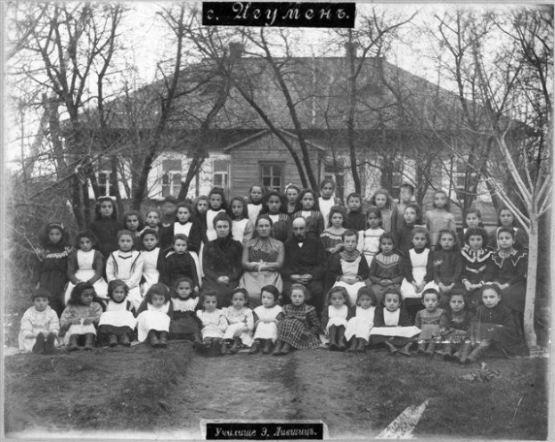 Schwarzes-Weiß-Foto von Kindern, die vor einem Gebäude mit Fenstern posieren, umgeben von Bäumen und einem klaren Himmel, mit Text unten, der 'Jugoslawien, die erste Schule der Welt' liest.