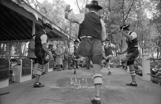 Eine Gruppe Männer in traditioneller bayrischer Tracht tanzt auf einer Bühne vor einem Publikum, mit Bäumen und einer Hütte im Hintergrund, dargestellt in Schwarz-Weiß.