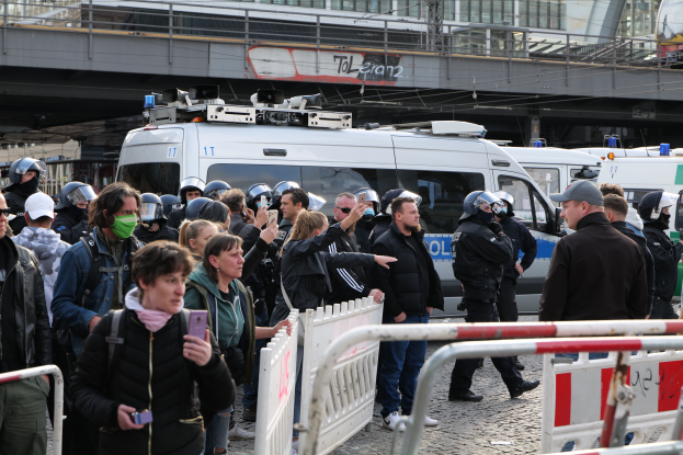 Eine Gruppe von Menschen steht vor einer Reihe von Polizeiwagen, einige tragen Helme und halten Telefone, mit Absperrungen im Vordergrund und einer Brücke mit Geländern und Gebäuden im Hintergrund, in Berlin, Deutschland.
