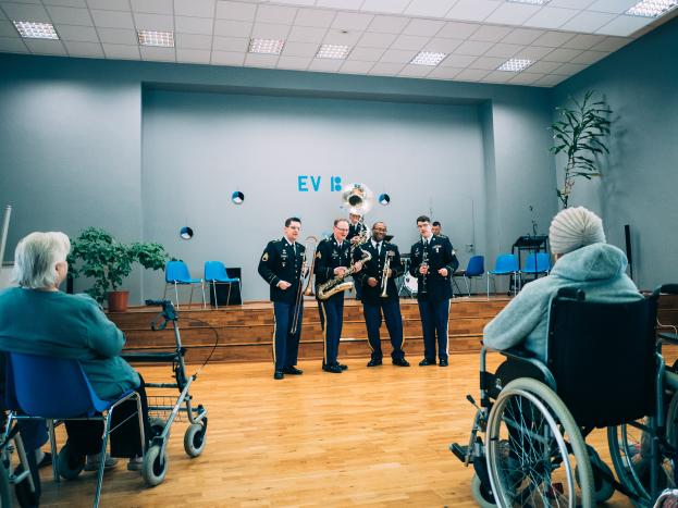 Eine Gruppe von Menschen im Rollstuhl, die in einem Raum mit Stühlen, Pflanzen und Deckenleuchten Musikinstrumente spielen, mit einigen Personen, die auf Stühlen sitzen, was auf ein Rollstuhlband-Event hindeutet.