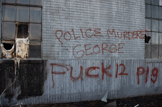 Graffiti an einem Gebäude mit der Aufschrift 'Polizei ermordet George Fuck 12 P19' und zerschlagenen Fenstern und verstreuten Gegenständen davor.