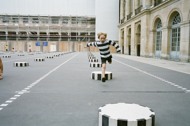 Ein Spielbereich mit vier Kindern, eines rennt auf dem Boden mit schwarzen und weißen Betonstufen, umgeben von einem Schulgeb├Ąude.