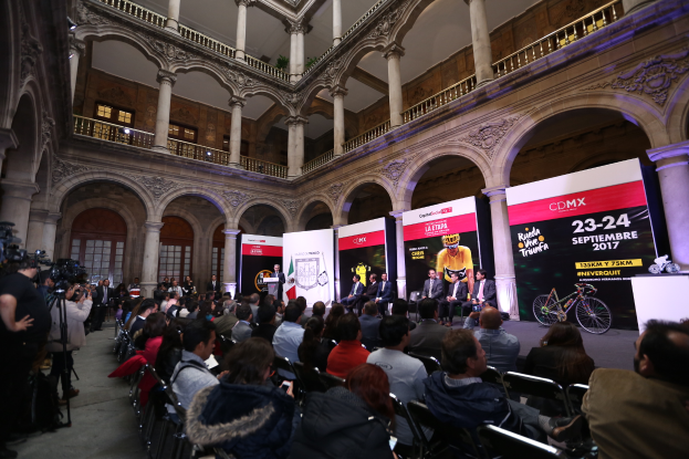 Eine Gruppe von Menschen sitzt an einem Tisch mit Mikrofon und Podium vor einer Menge, Kameras und Bannern, was auf eine Pressekonferenz für das CDMX 2017-Event hinweist.