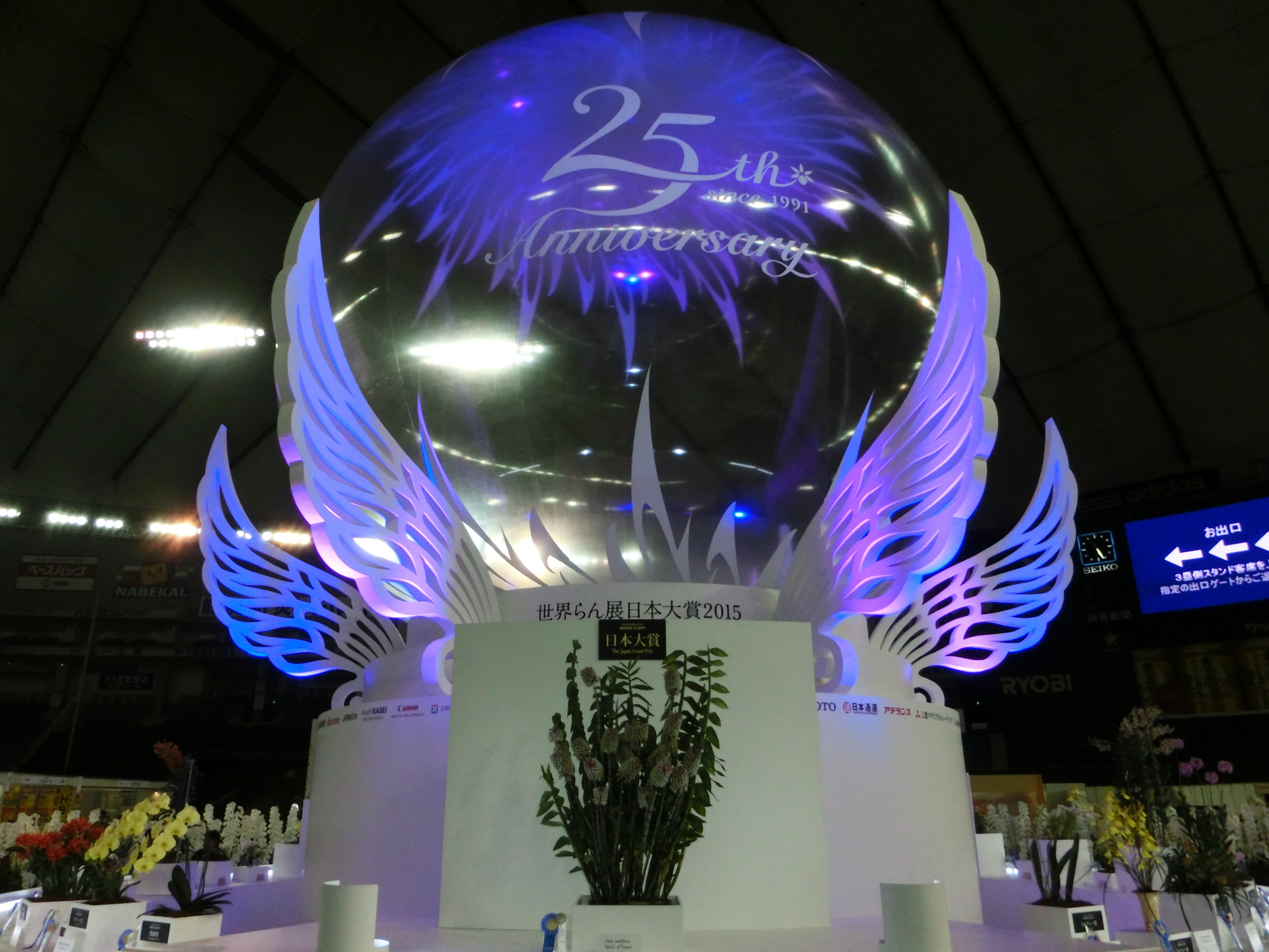 Ein großer Kristallball mit Flügeln oben drauf, umgeben von Blumenschmuck, Namensschildern und anderen Gegenständen auf dem Boden, vor einer Kulisse aus Plakaten, Lichtern und einem Bildschirm während einer 25-jährigen Jubiläumsfeier.